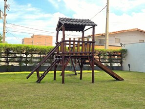 A wooden play structure with a roof and slides in a grassy yard. - Amaranth By Hosppedar (Cabedelo)