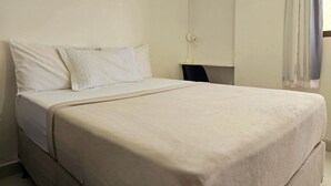 Una cama con una colcha blanca y almohadas, una mesita de noche con una lámpara y una ventana con cortinas.