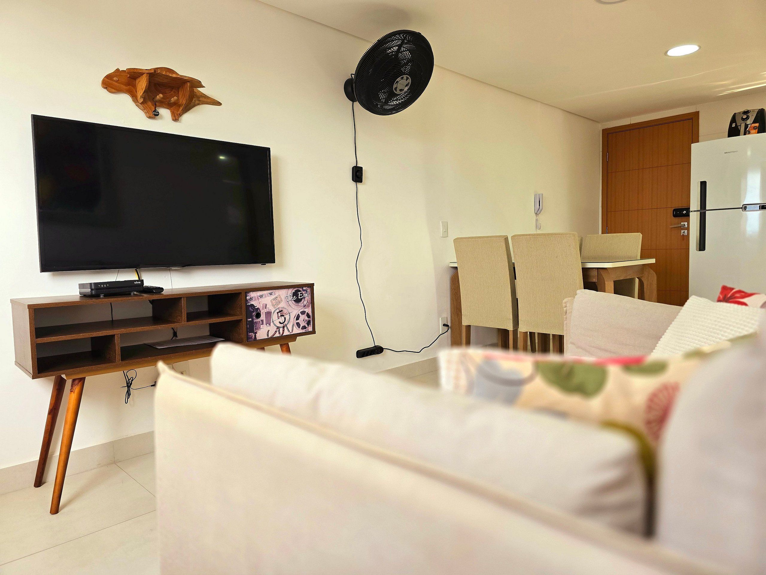 Una sala de estar con un televisor de pantalla plana, un mueble de madera para equipo audiovisual, un ventilador de pie y un comedor con mesa y sillas.