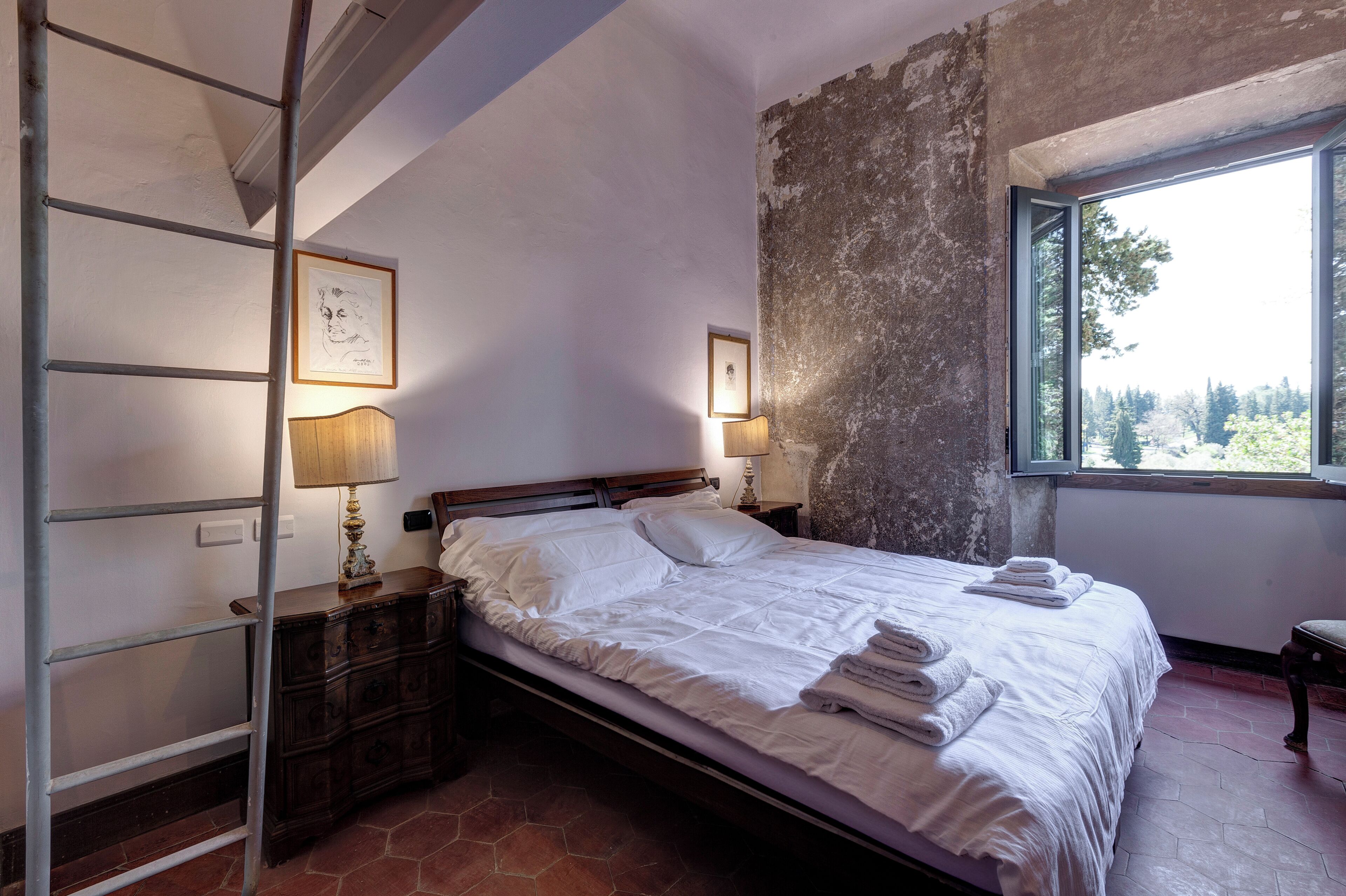 Une chambre à coucher avec un lit, deux lampes de chevet, une table de nuit en bois, une commode et une fenêtre donnant sur des arbres.