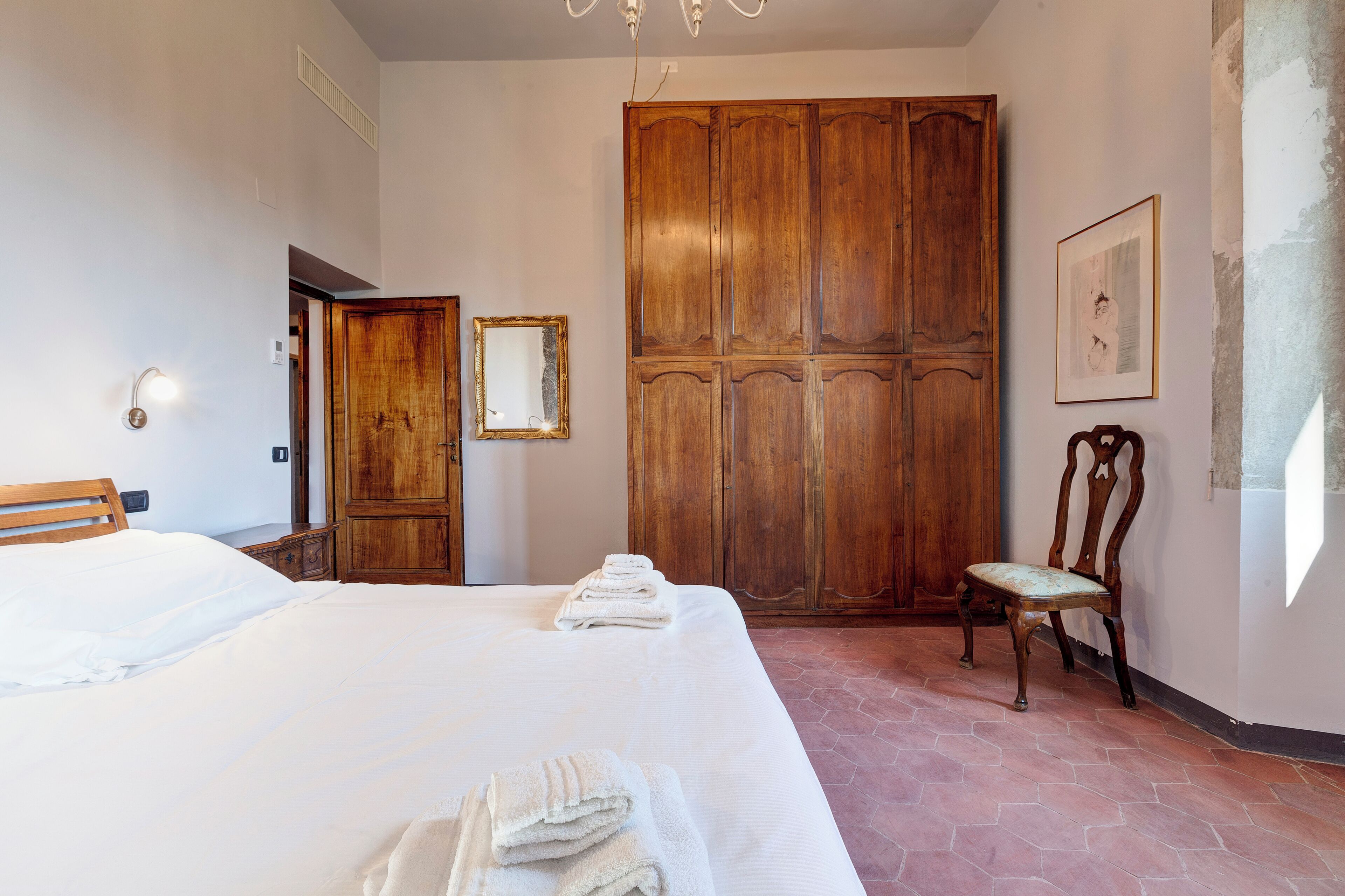 A hotel room with a large bed, a wooden wardrobe, a chair, and a framed picture on the wall.