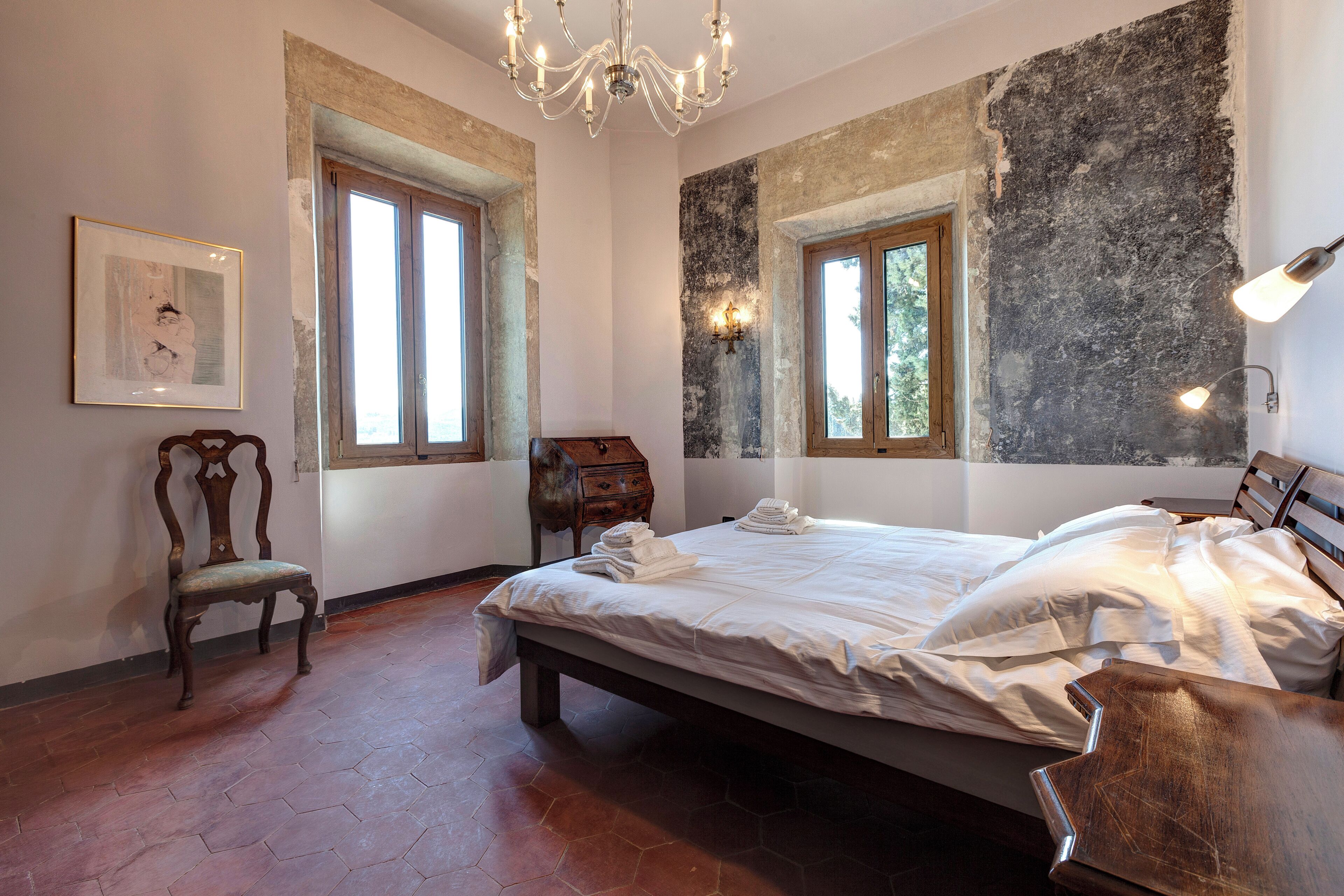 Une chambre à coucher avec un grand lit, deux fenêtres, un lustre et une chaise en bois.
