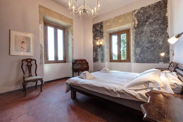 Un dormitorio con una cama grande, dos ventanas, una araña y una silla de madera.