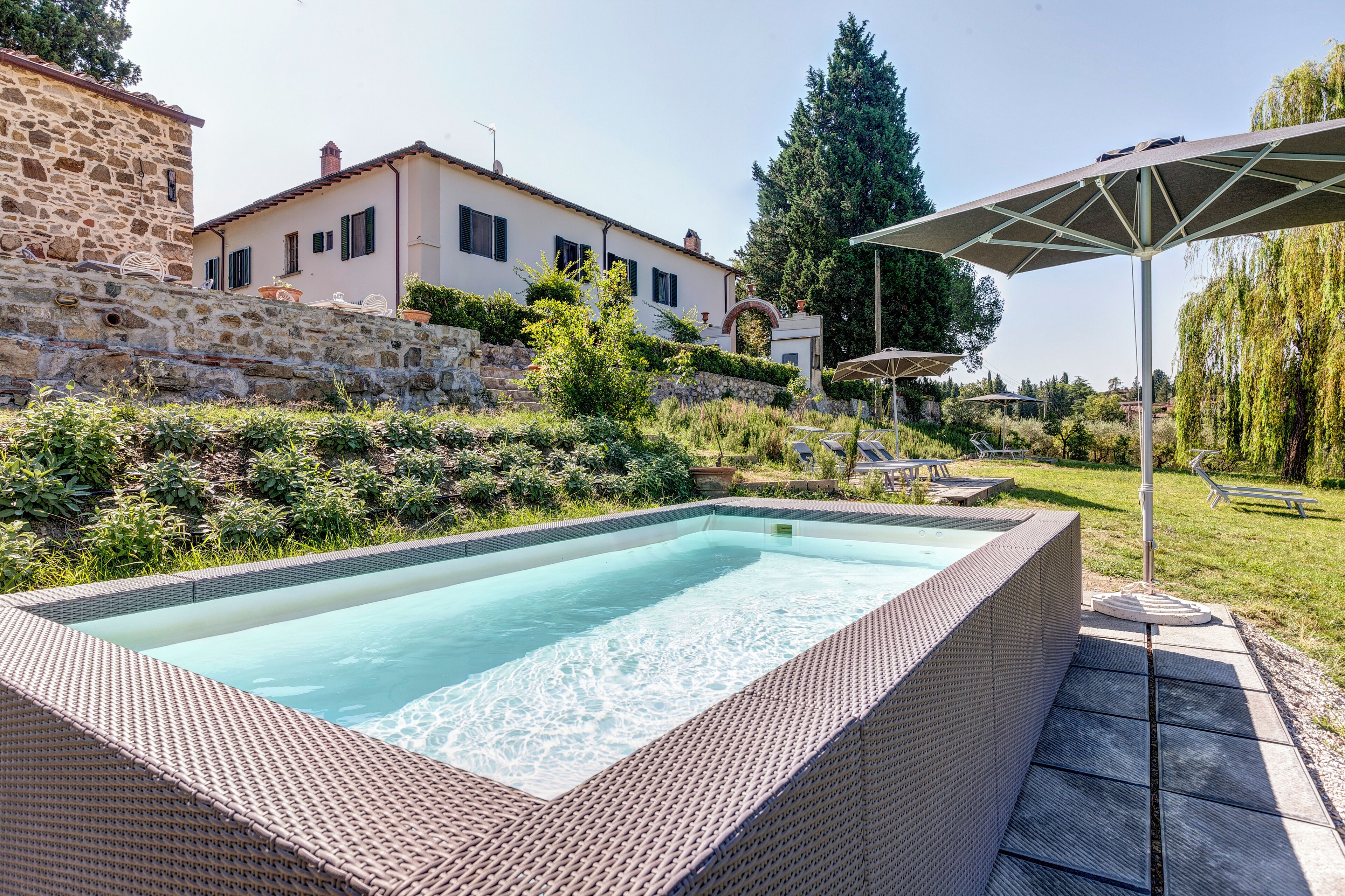 A rectangular swimming pool with a wicker edge, surrounded by a stone wall and lush greenery.