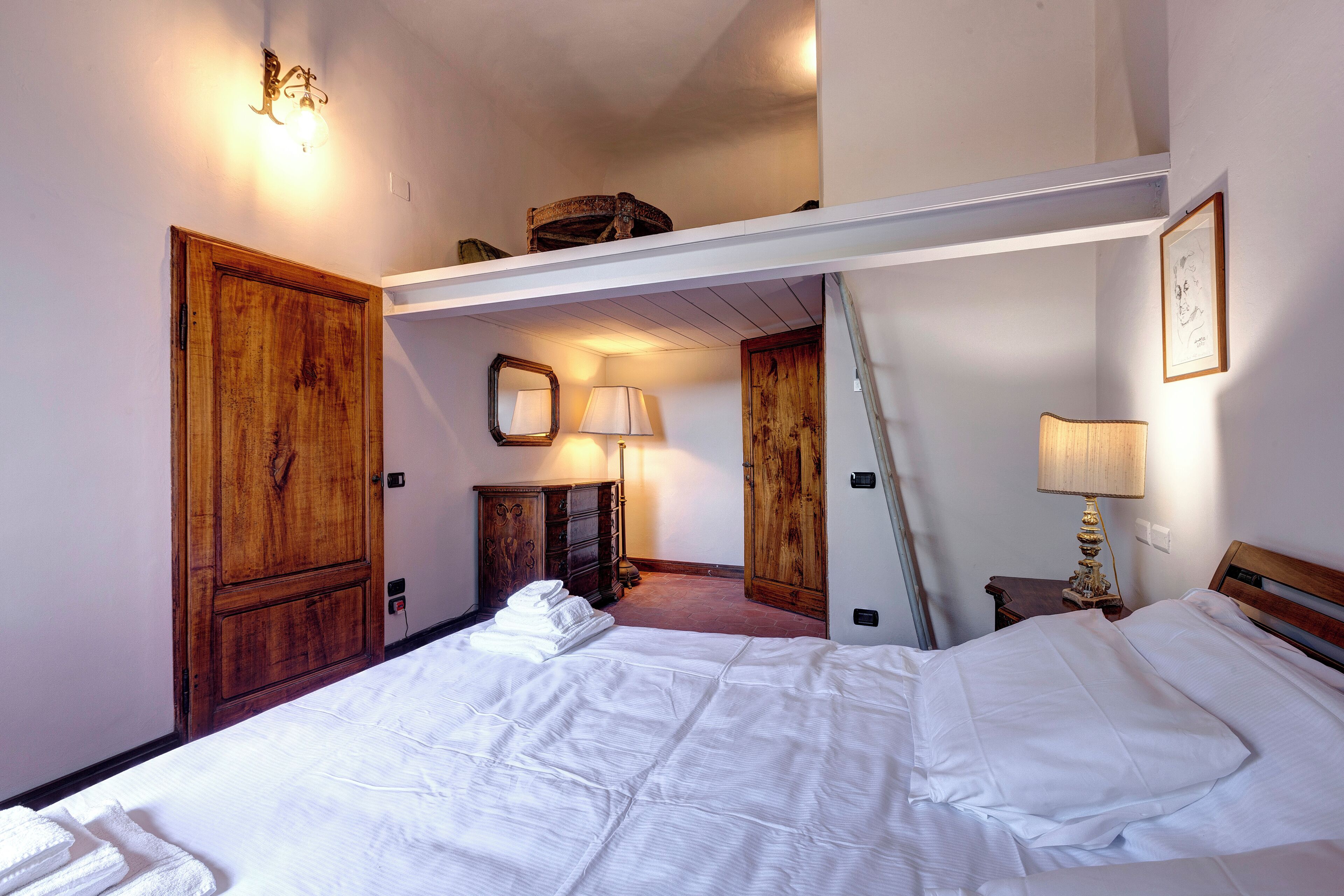 A hotel room with a bed, a wooden wardrobe, a mirror, a lamp, and a picture on the wall.