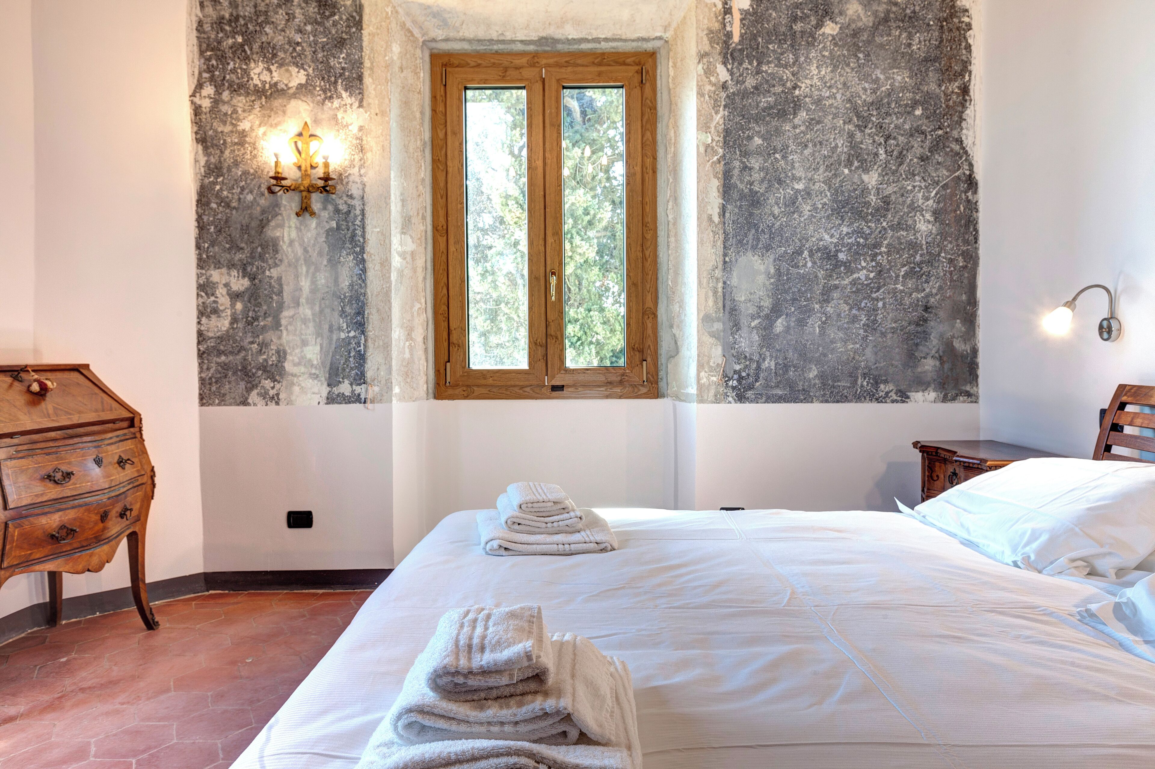 A bedroom with a large bed, a wooden dresser, a window with a view of trees, and a wall-mounted light fixture.