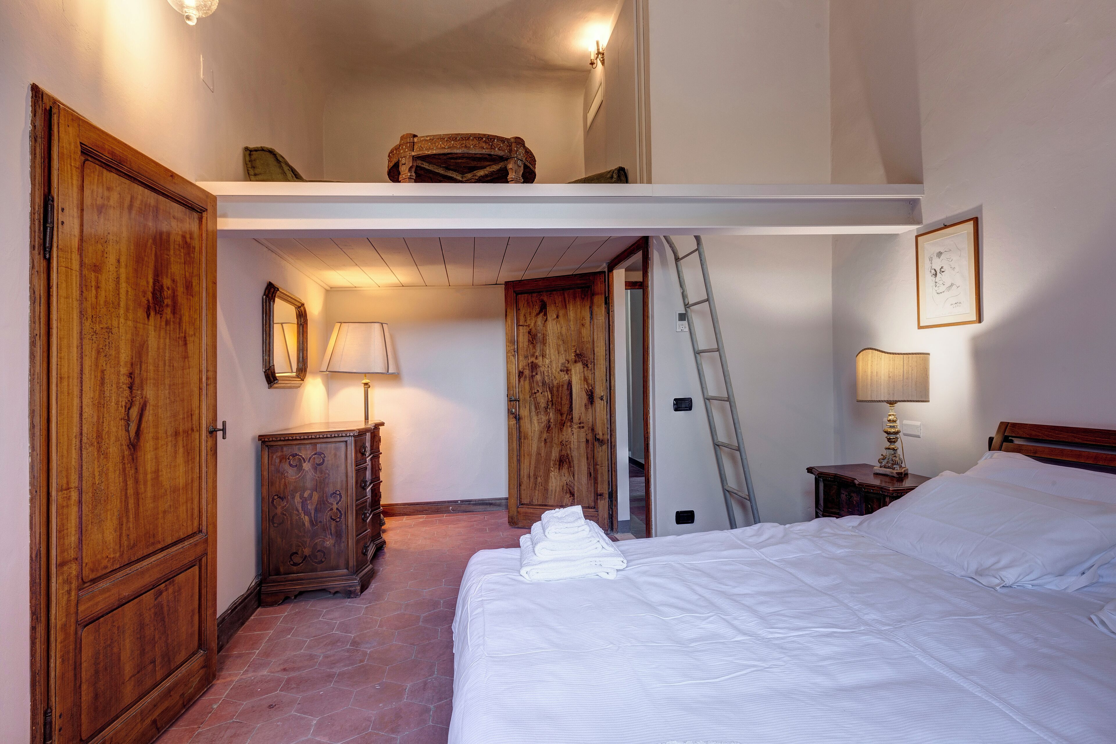 A bedroom with a large bed, wooden wardrobe, a dresser, a desk, a ladder, and a door leading to another room.
