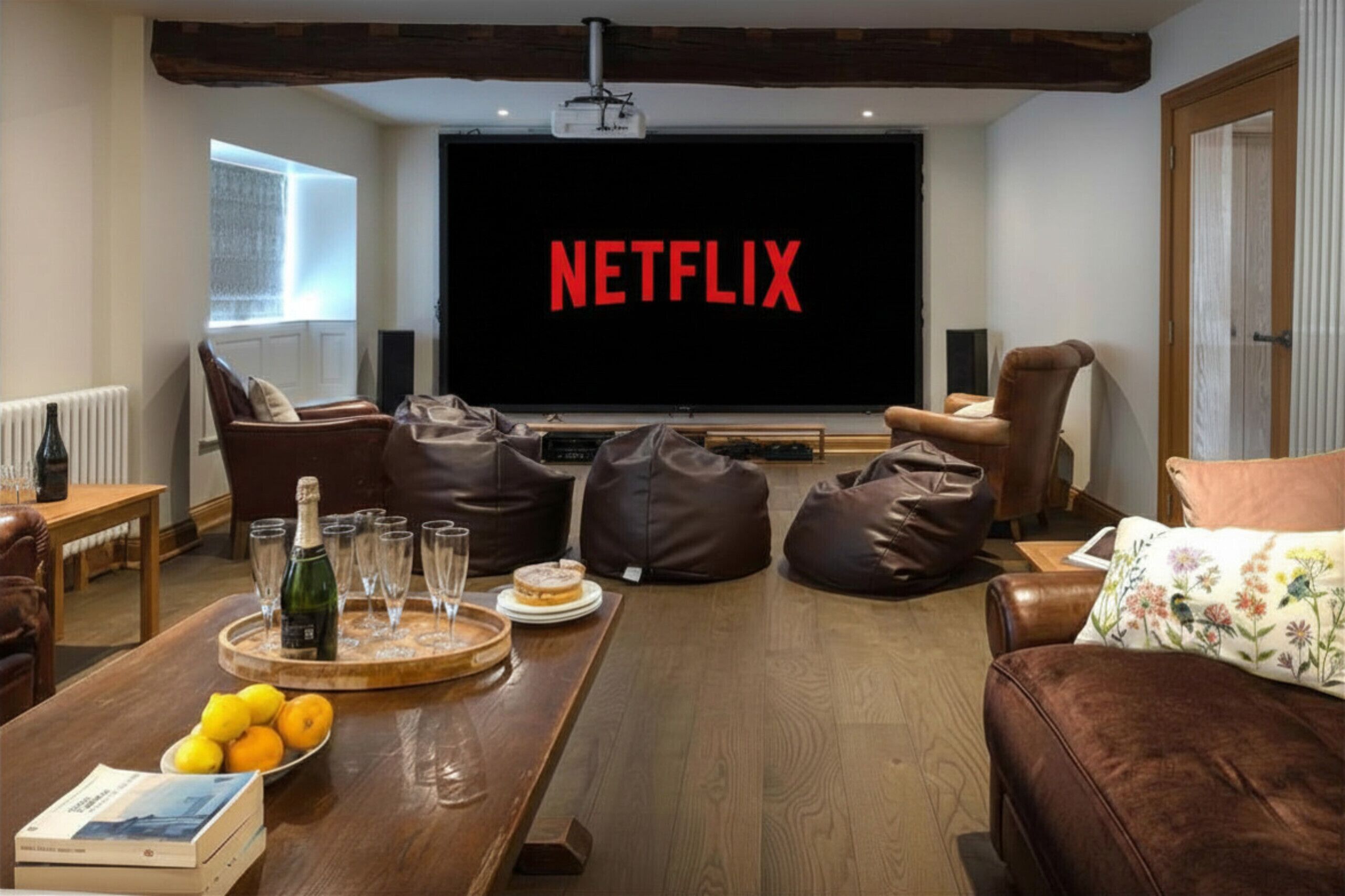 Una sala de estar con una pantalla grande mostrando 'NETFLIX', sofás de cuero, una mesa de centro de madera con bebidas y fruta, y un proyector.
