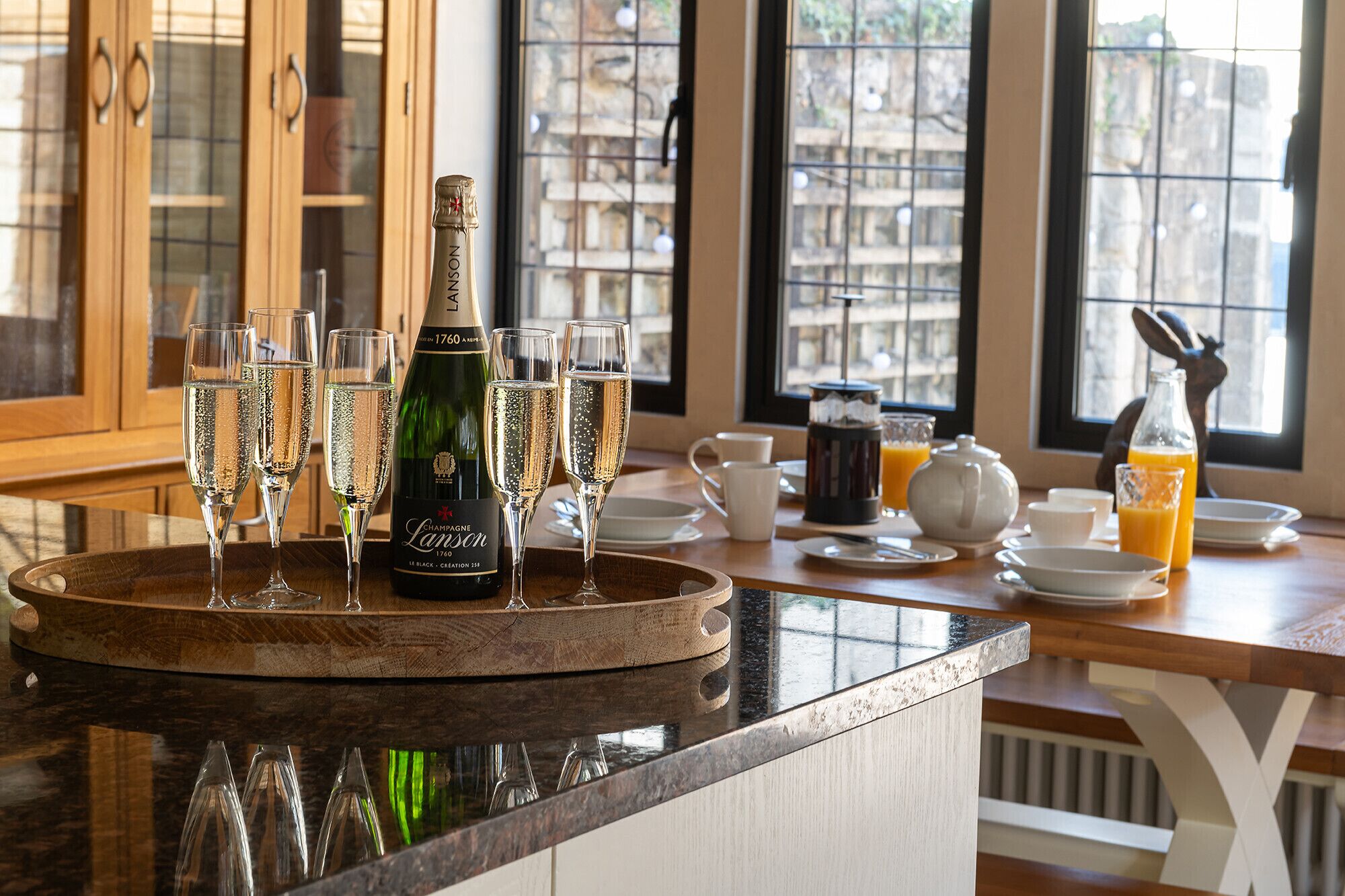 Une table de petit-déjeuner avec du champagne, des verres, une cafetière et du jus d’orange, le tout disposé sur un plateau en bois.