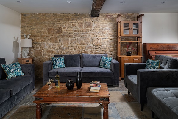 A living room with a stone wall, a dark grey sofa, matching cushions, a wooden coffee table with books and a bottle, and a wooden cabinet with glass doors.