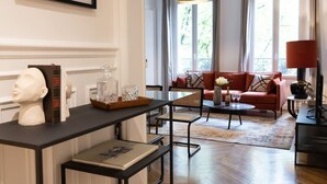 A living room with a sofa, a coffee table, and a television. There is a window with curtains and a chandelier. - A2-8 people-4 bedrooms-Wagram-AC-Paris 8 (Paris)