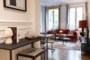 A living room with a sofa, a coffee table, and a television. There is a window with curtains and a chandelier. - A2-8 people-4 bedrooms-Wagram-AC-Paris 8 (Paris)
