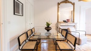 A dining area with a glass table surrounded by wicker chairs, a large mirror, and a vase with flowers. - A2-8 people-4 bedrooms-Wagram-AC-Paris 8 (Paris)