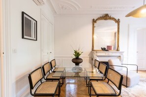 A dining area with a glass table surrounded by wicker chairs, a large mirror, and a vase with flowers. - A2-8 people-4 bedrooms-Wagram-AC-Paris 8 (Paris)