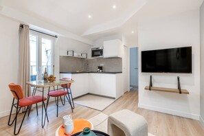 Ein modernes Wohnzimmer mit Flachbildfernseher, einem Essbereich mit Glastisch und einer Küche mit weißen Schränken.