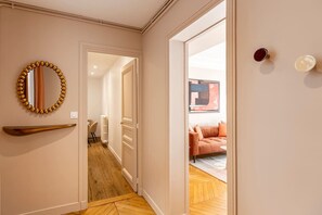 Um corredor com piso de madeira, um espelho com moldura dourada e uma porta que dá para um quarto com sofá e quadro.
