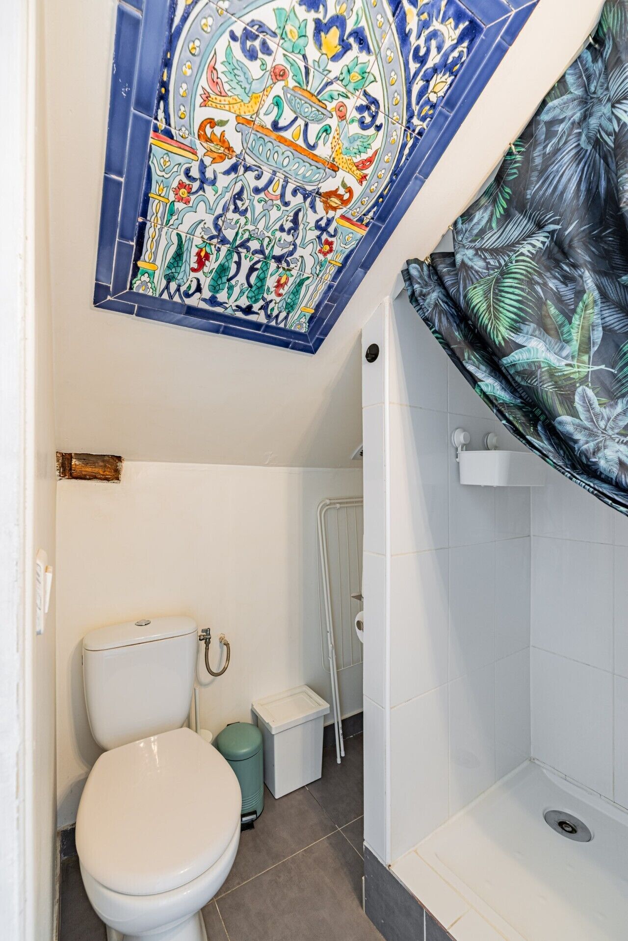 A bathroom with a white toilet, a trash bin, a shower area, and a decorative tile ceiling.