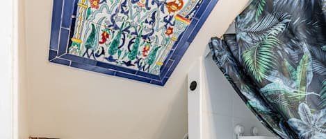 A bathroom with a white toilet, a trash bin, a shower area, and a decorative tile ceiling.