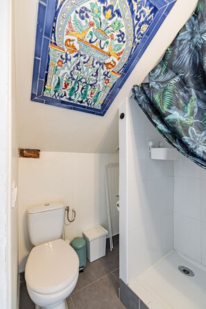 A bathroom with a white toilet, a trash bin, a shower area, and a decorative tile ceiling. - Palais Royal – Louvre Museum – Central Studio (Paris)