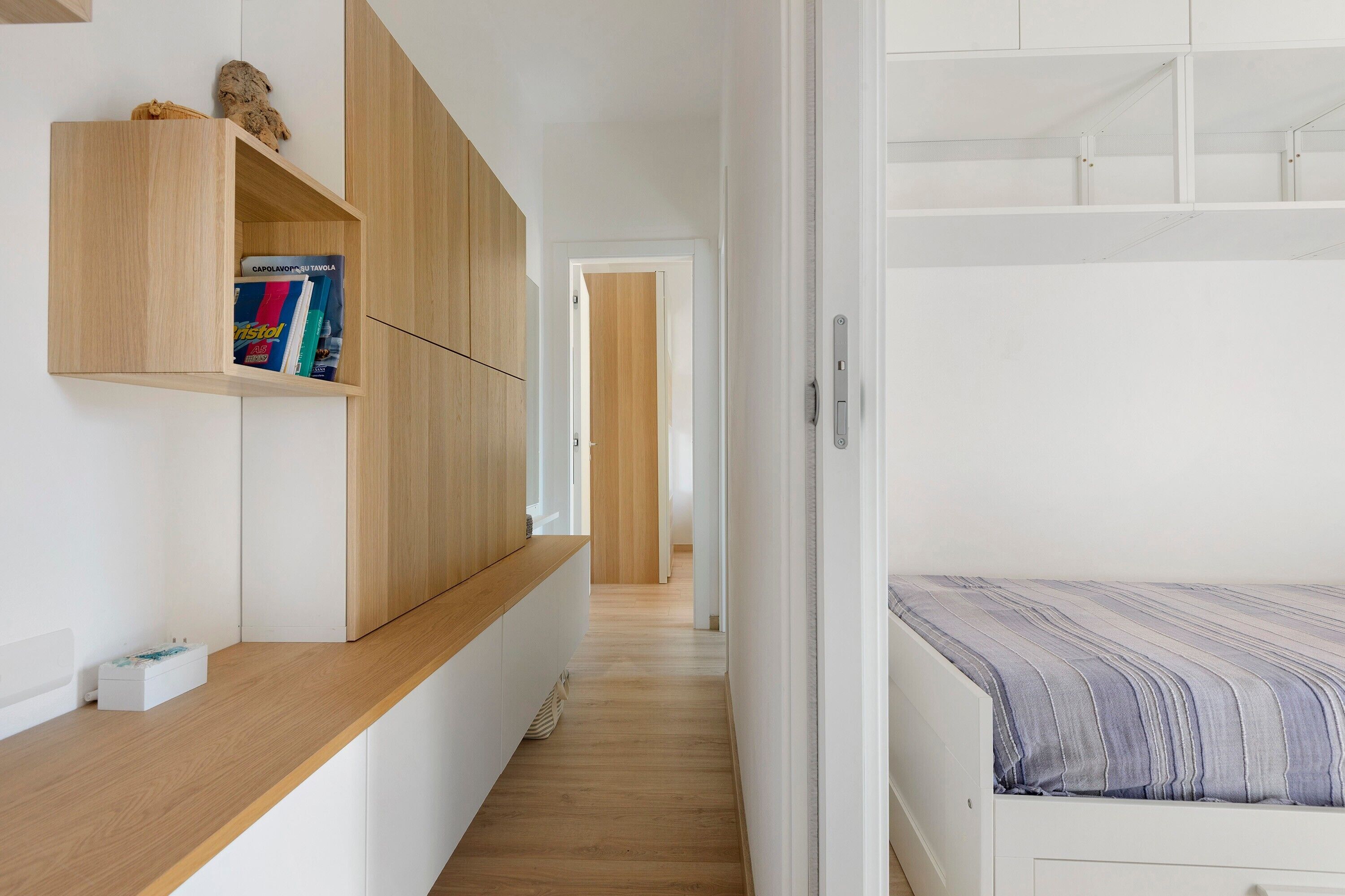 Un couloir étroit avec un banc en bois et un lit recouvert d’une couverture à rayures.