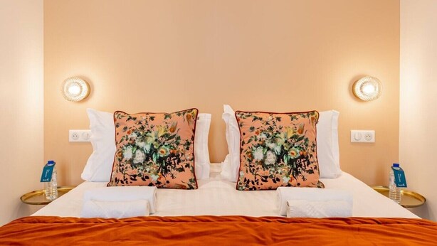 Une chambre d’hôtel avec un lit, deux oreillers ornés de motifs floraux, des tables de chevet avec des bouteilles d’eau et des lampes fixées au mur.