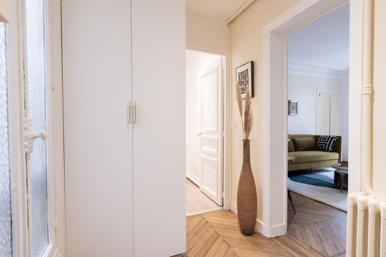 Ein Flur mit einer weißen Tür, einer Vase mit Pampasgras und einem grünen Sofa im Hintergrund.