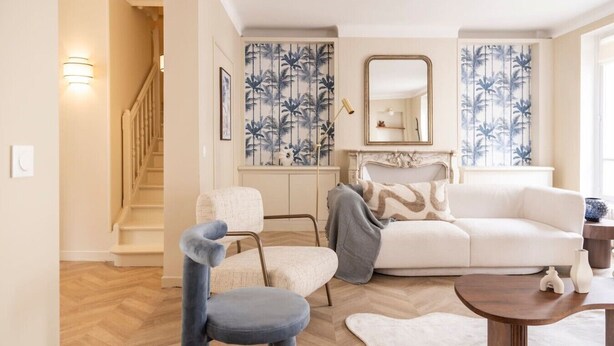 Una sala de estar con un sofá blanco, una silla azul, una mesa de centro de madera y una pared con diseño.