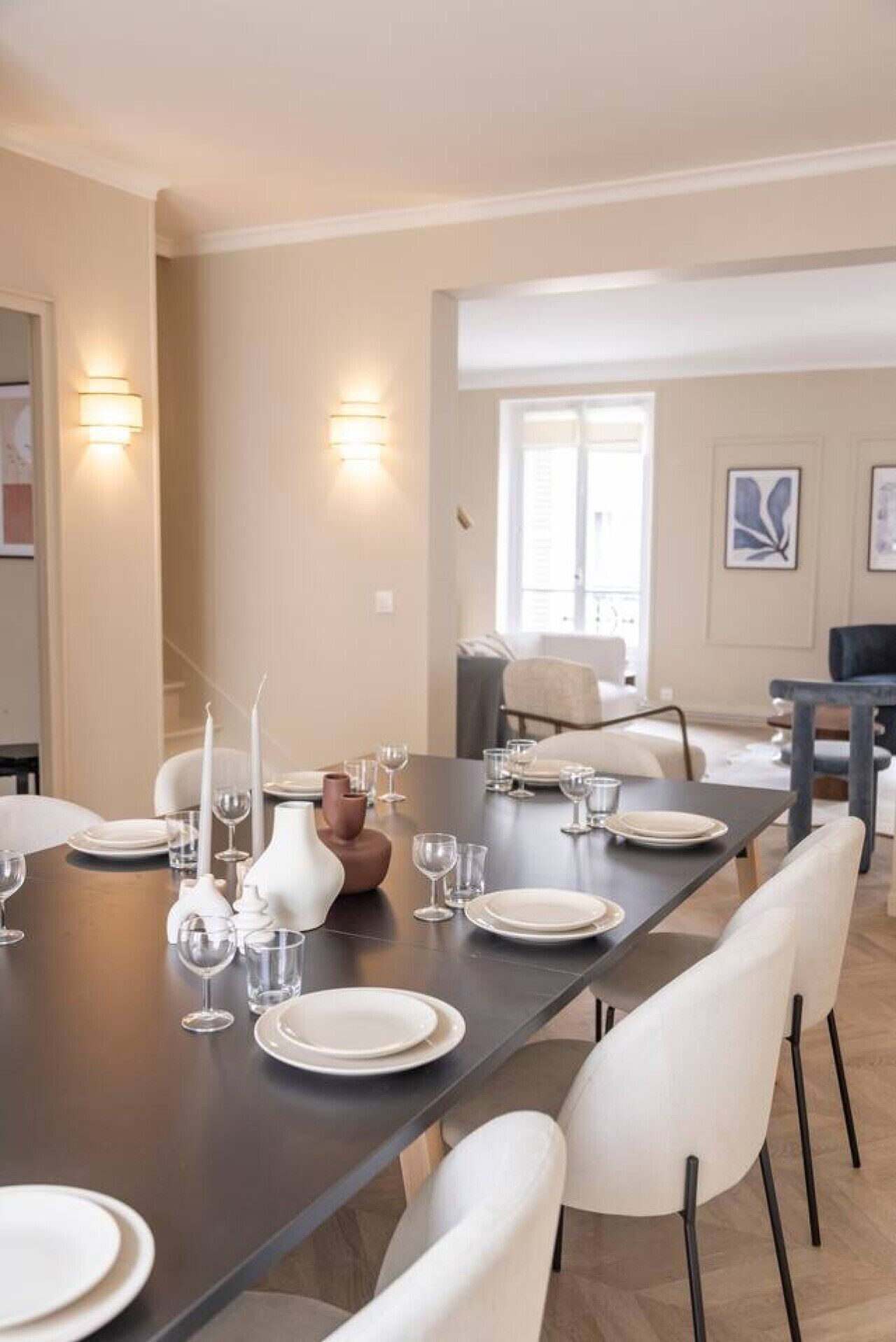 A dining area with a long table set for a meal, chairs, wall-mounted light fixtures, and a window with blinds.