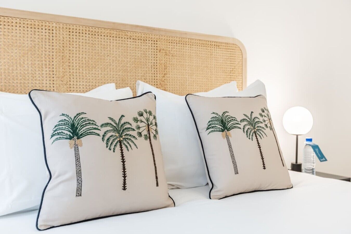 A bed with two pillows featuring palm tree designs, a round bedside table with a water bottle, and a wall-mounted lamp.