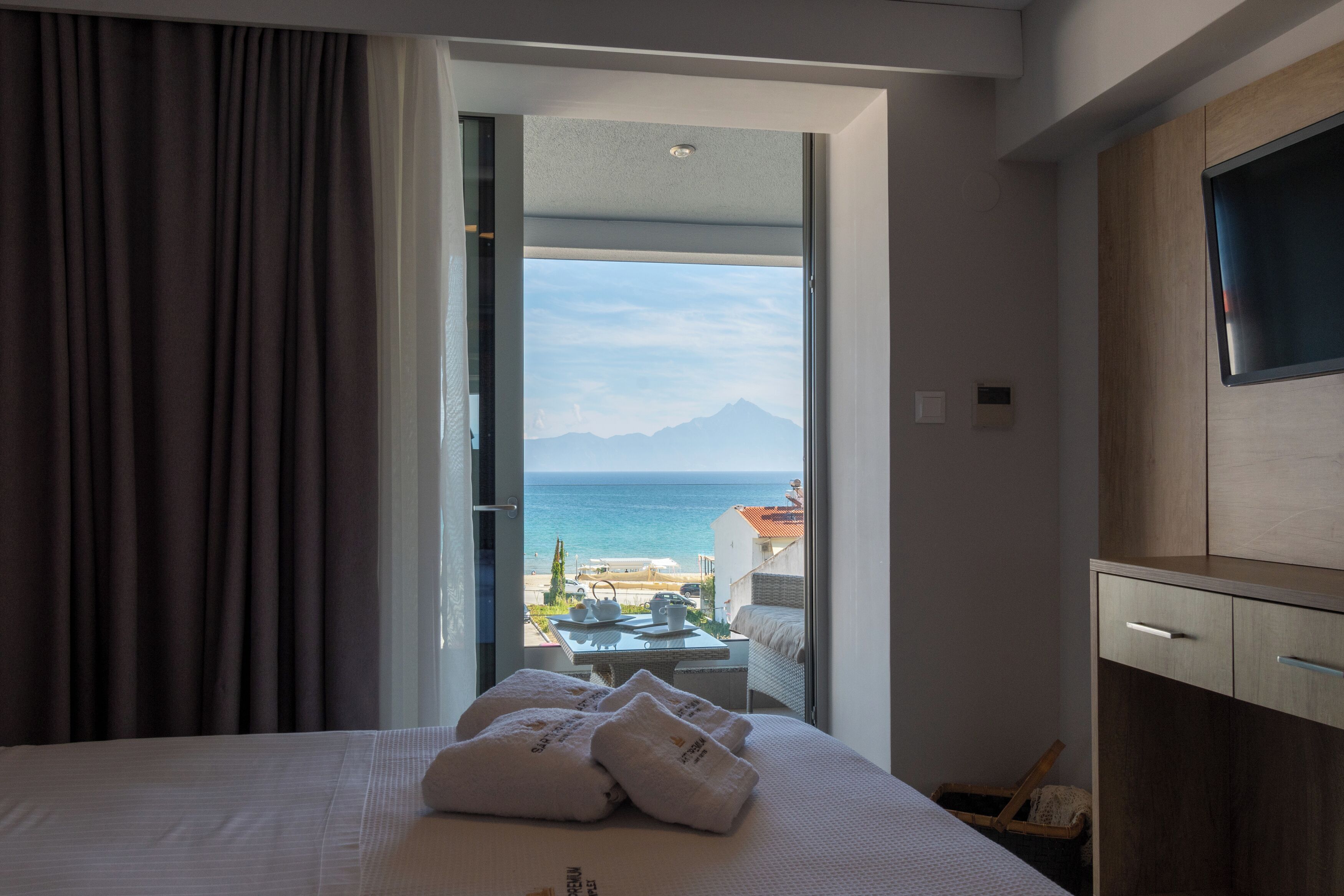 A hotel room with a bed, a TV, a desk, and a view of the sea and mountains.