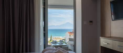 A hotel room with a bed, a TV, a desk, and a view of the sea and mountains.