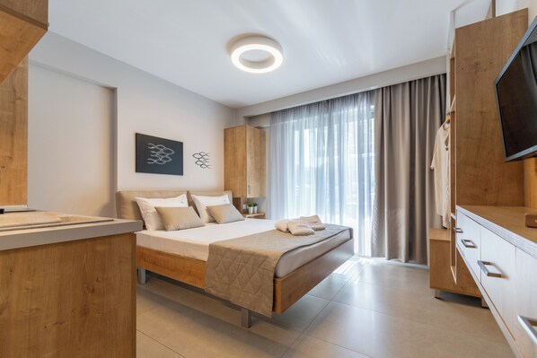 A modern hotel room with a bed, wooden furniture, a flat-screen TV, and a large window with curtains. - SARTI PREMIUM LUXURY SUITES (SITHONIA)