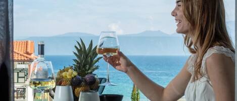 A person seated at a table with a glass of drink, a bottle of wine, and a bowl of fruit. The table is set with plates and cutlery. In the background, there is a view of the sea and mountains.