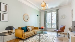 Una sala moderna con un sofá, una mesa de centro y una silla. Hay una ventana con cortinas, un espejo en la pared y una lámpara.