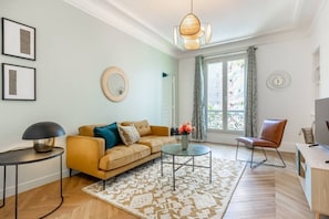 Ein modernes Wohnzimmer mit einer Couch, einem Couchtisch und einem Sessel. Ein Fenster mit Vorhängen, ein wandmontierter Spiegel und eine Lampe sind ebenfalls vorhanden.
