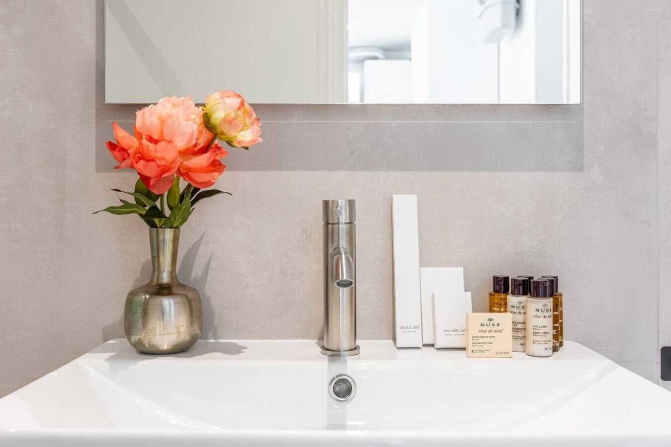 A bathroom with a white sink, a vase with flowers, a mirror, and toiletries.
