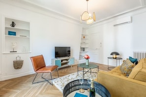 Ein modernes Wohnzimmer mit Fernseher, einem braunen Ledersessel, einem runden Couchtisch mit Weingläsern und einem Sofa mit gemustertem Überwurf.