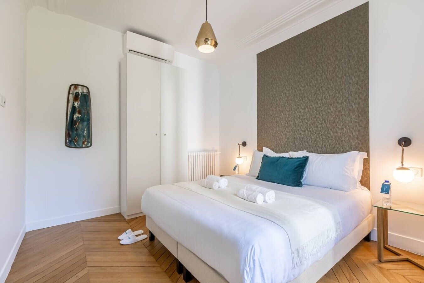 Une chambre d’hôtel avec un lit, des tables de chevet, un radiateur, un luminaire suspendu et une porte.