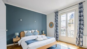 A bedroom with a bed, wooden headboard, two bedside tables, a blue rug, a window with curtains, and a view of buildings outside. - Heart of Paris, 2-Bedroom Apartment, Arts et Métiers, Marais (Paris)