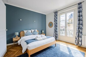 A bedroom with a bed, wooden headboard, two bedside tables, a blue rug, a window with curtains, and a view of buildings outside. - Heart of Paris, 2-Bedroom Apartment, Arts et Métiers, Marais (Paris)
