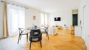 A modern living room with a round dining table, black chairs, a white sofa, and a flat-screen TV mounted on the wall. - Heart of Paris, 2-Bedroom Apartment, Arts et Métiers, Marais (Paris)
