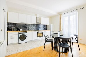 A modern kitchen with white cabinets, a dishwasher, an oven, and a dining area with a round table and chairs. - Heart of Paris, 2-Bedroom Apartment, Arts et Métiers, Marais (Paris)