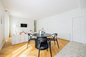 A modern living room with a round table, black chairs, a white sofa, and a television mounted on the wall. - Heart of Paris, 2-Bedroom Apartment, Arts et Métiers, Marais (Paris)