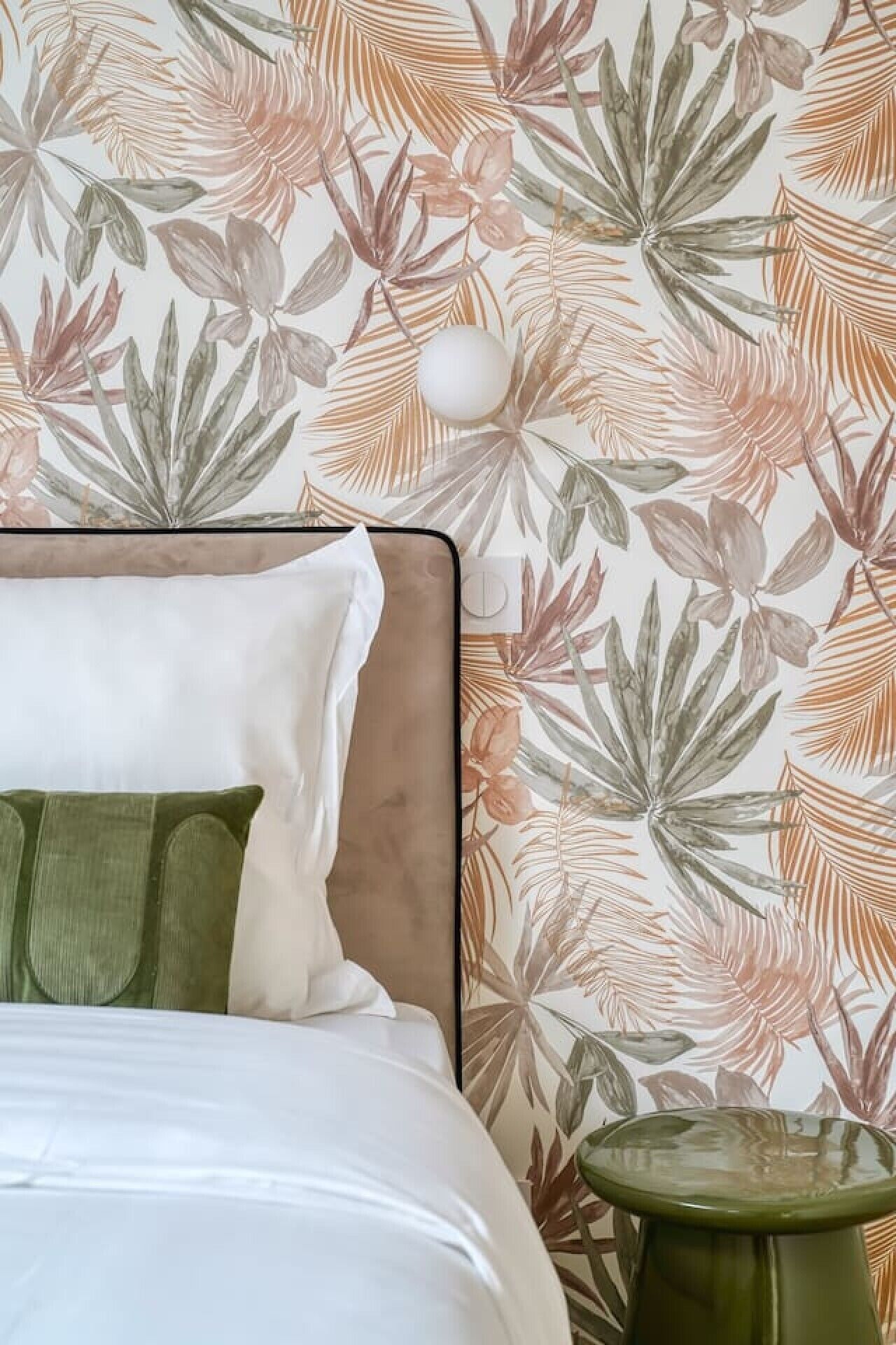A bed with white bedding and green pillows, a headboard, and a wall with a tropical leaf pattern.