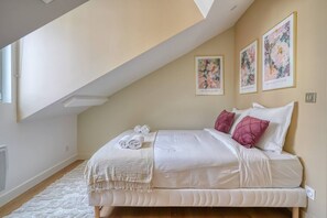 Quarto com teto inclinado, cama com roupa de cama branca e almofadas cor-de-rosa, manta branca e duas obras de arte florais emolduradas na parede.