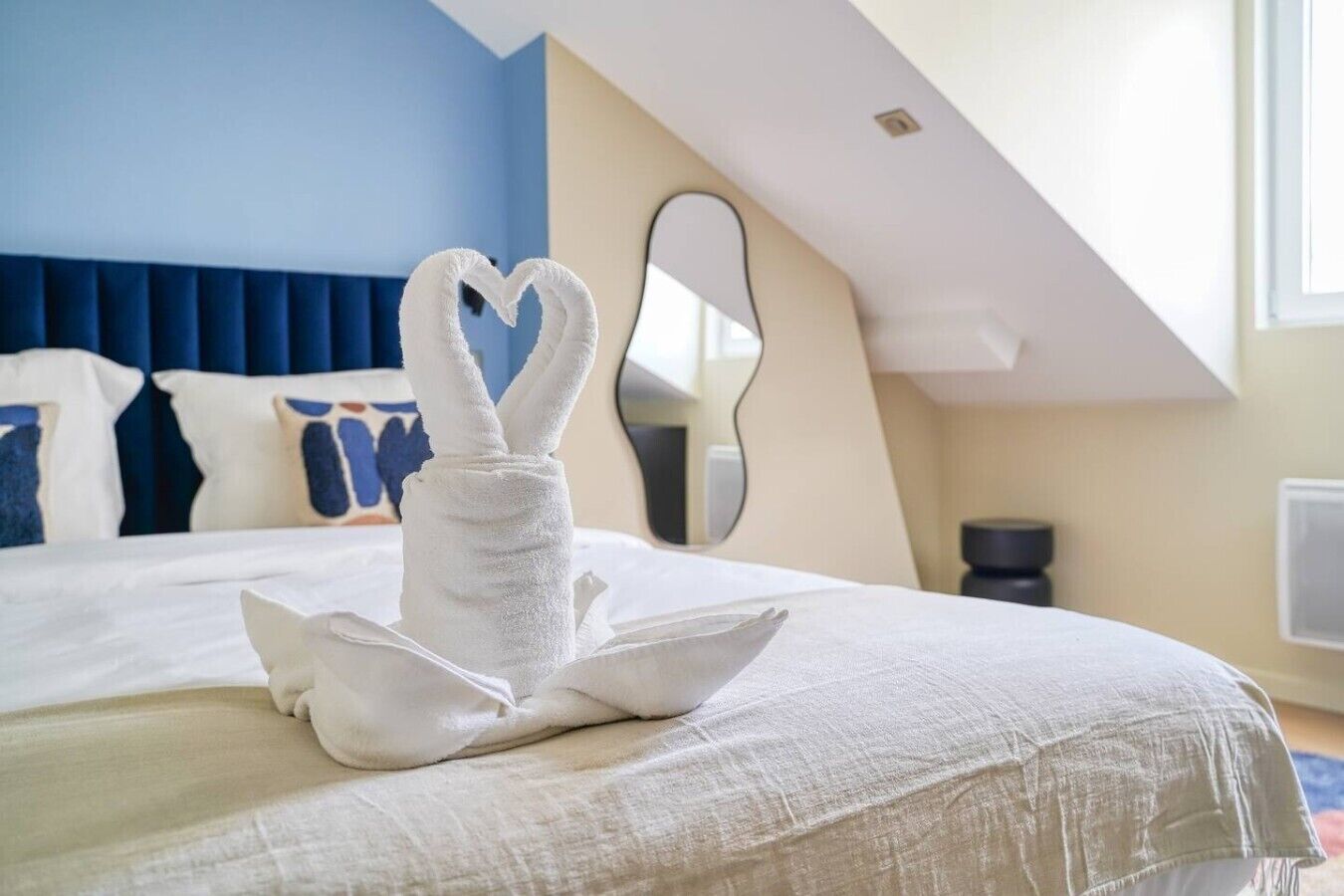 A hotel room with a bed, a heart-shaped towel decoration, a mirror, and a blue wall.