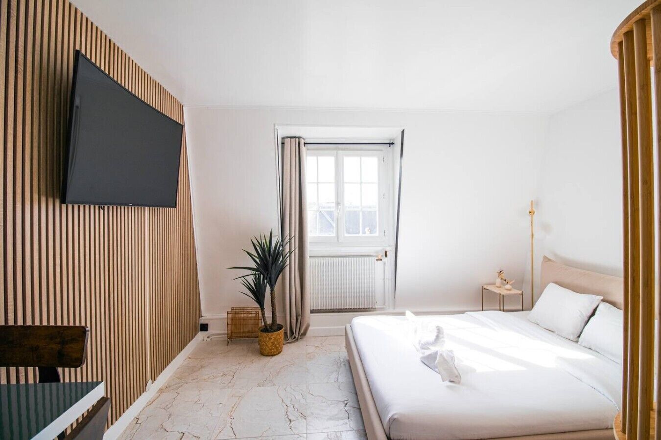 A hotel room with a bed, a television, a window with curtains, a plant, and a radiator.