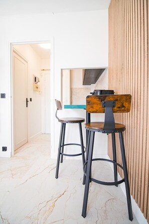 A kitchen area with a wooden chair and a bar stool, a countertop, and a wall-mounted television. - Studio for 2 People - Hotel de Ville - Marais - Paris 04 - Central (Paris)