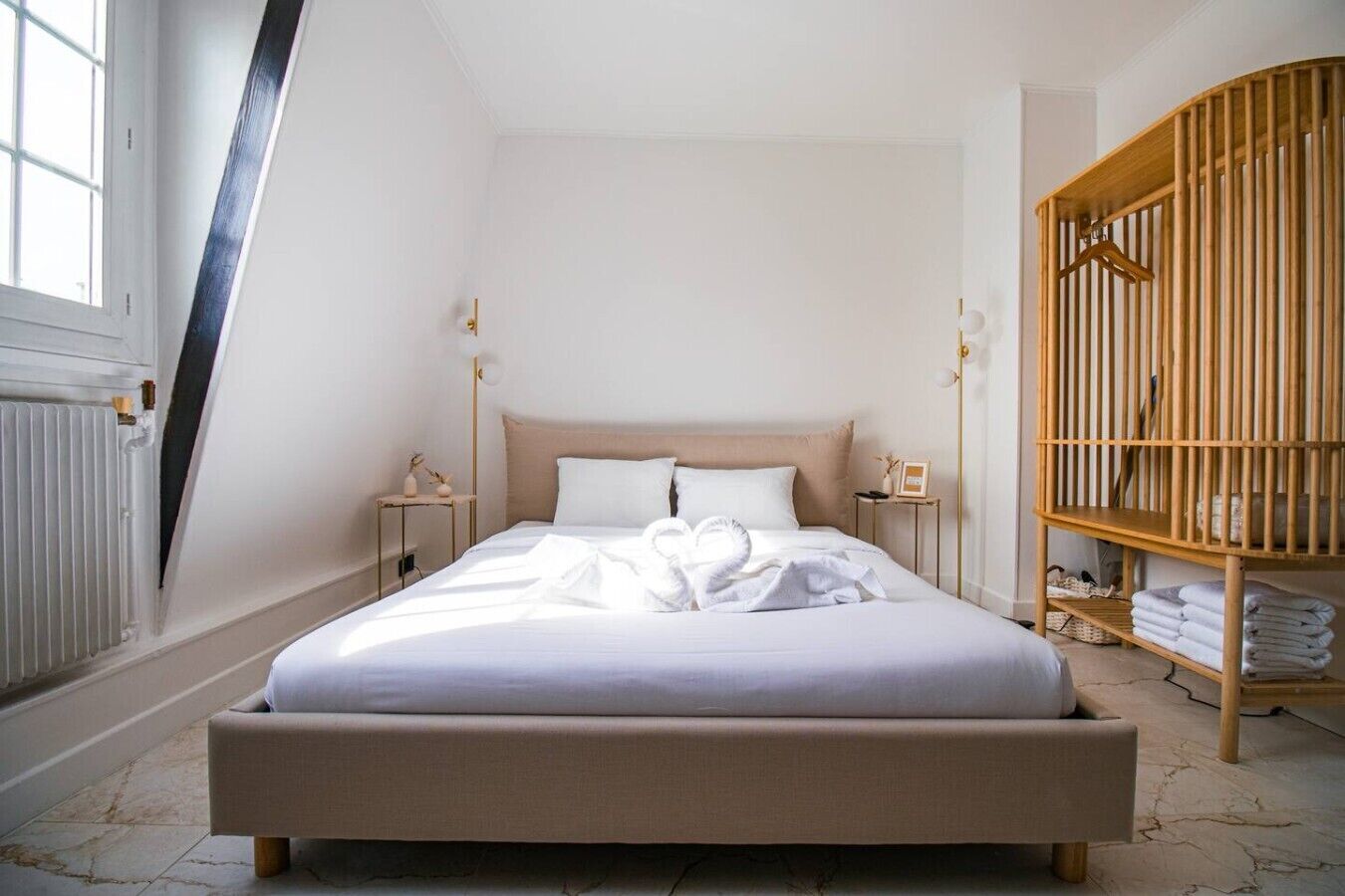 Un lit bien fait, avec du linge de lit blanc, dans une pièce dont le plafond est en pente, avec une armoire en bois et une petite table de chevet surmontée d’une lampe.