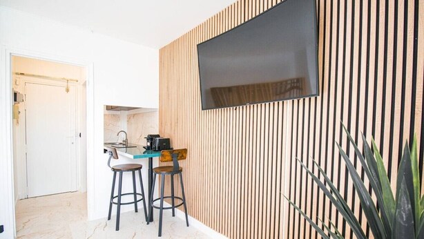 A modern kitchen with a built-in stove, a dining area with two stools, and a large flat-screen TV mounted on a wooden slatted wall. - Studio for 2 People - Hotel de Ville - Marais - Paris 04 - Central (Paris)